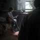 Joel stands in the threshold of a room watching on as Ellie strums her guitar on a bed that has blood splatter and a knife in front of it.